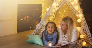 Mother and daughter reading a book together under a festive tent, illuminated by warm lights near a propane-powered fireplace.